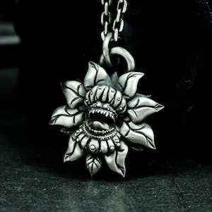 Könnte beinhalten: Eine silberne Halskette mit einem Anhänger, der ein detailliertes, verziertes Blumendesign mit einem aufgerissenen Mund und scharfen Zähnen zeigt. Die Blume hat eine strukturierte, dreidimensionale Optik.