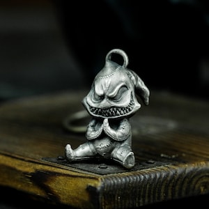 Könnte beinhalten: Ein silberfarbener Schlüsselanhänger aus Metall in Form eines kleinen, cartoonartigen Monsters mit einem breiten Grinsen und großen Augen. Das Monster sitzt mit den Händen vor sich gefaltet.