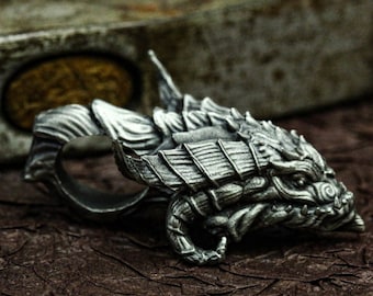 May include: A silver dragon head ring with intricate details. The ring is made of metal and has a textured surface. The dragon's head is facing to the right and has a fierce expression.