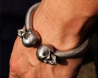May include: A silver-toned metal cuff bracelet featuring two skull accents. The bracelet has a curved design that wraps around the wrist. The skulls are detailed with eye sockets, noses, and teeth.