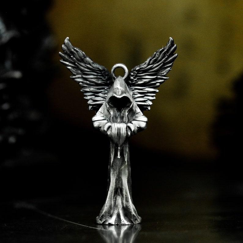 May include: A silver pendant featuring a hooded angel figure with outstretched wings. The angel is wearing a long robe and has its hands clasped in front of its chest.