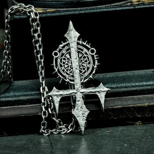 Może przedstawiać: Srebrny naszyjnik z zawieszką w kształcie krzyża z detalami i okrągłym symbolem z misternymi wzorami. Łańcuszek jest srebrny i ma splot typu ogniwa.