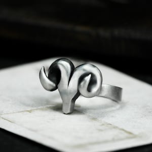 Może przedstawiać: Srebrny pierścionek z detalicznym wzorem głowy barana. Pierścionek jest wykonany z metalu i ma gładkie wykończenie.