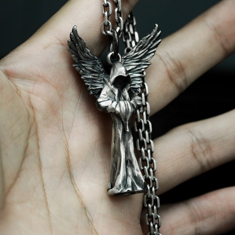 May include: A silver pendant necklace featuring a detailed sculpture of an angel with wings, wearing a hooded robe. The angel is standing with its arms crossed and has a serious expression.