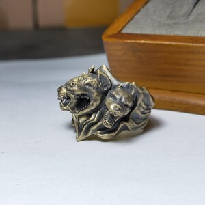 Puede incluir: Un anillo met&aacute;lico con un dise&ntilde;o esculpido de tres cabezas caninas gru&ntilde;endo con dientes expuestos y detalles de pelaje texturizado. El anillo tiene un acabado de lat&oacute;n antiguo con &aacute;reas hundidas oscurecidas para resaltar los rasgos faciales.