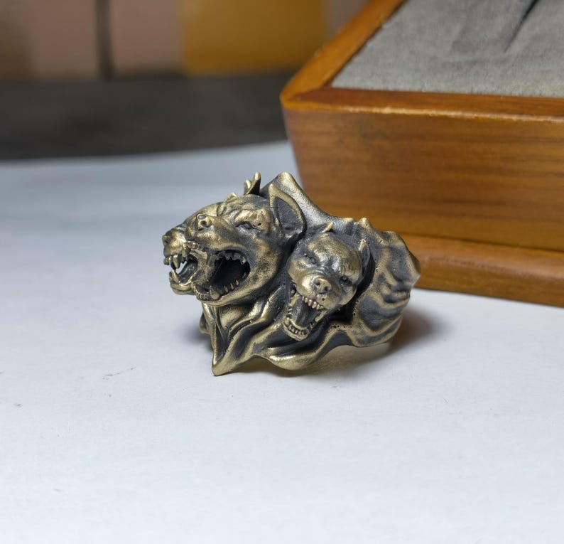 Puede incluir: Un anillo met&aacute;lico con un dise&ntilde;o esculpido de tres cabezas caninas gru&ntilde;endo con dientes expuestos y detalles de pelaje texturizado. El anillo tiene un acabado de lat&oacute;n antiguo con &aacute;reas hundidas oscurecidas para resaltar los rasgos faciales.