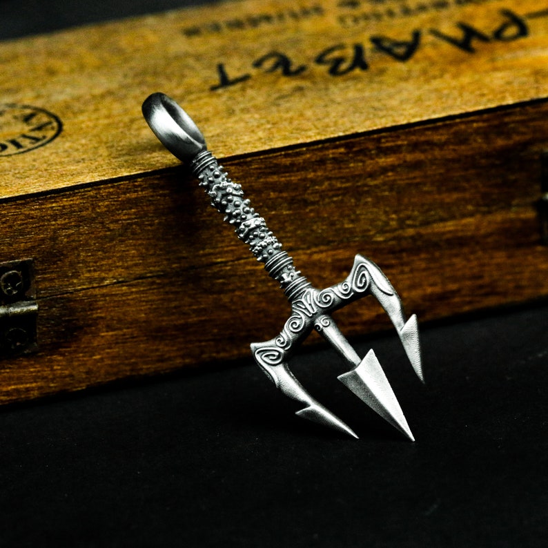 May include: A silver trident pendant with intricate details and a textured handle. The pendant is on a wooden surface.