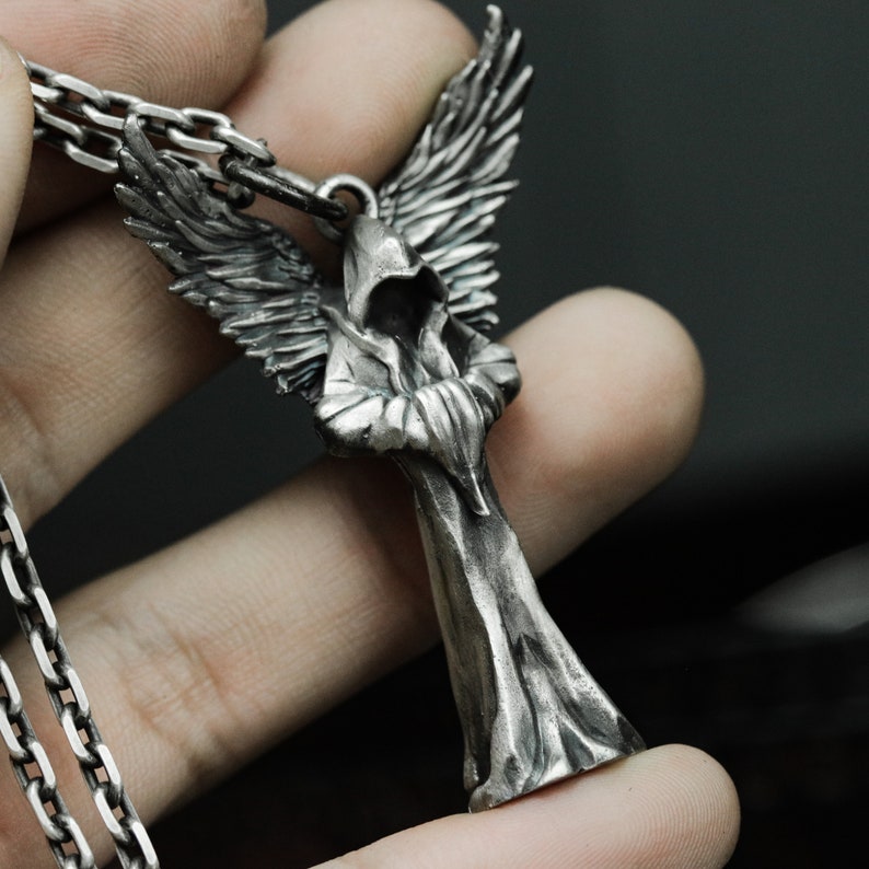 May include: Silver pendant necklace featuring a detailed sculpture of an angel wearing a hooded robe with wings outstretched.