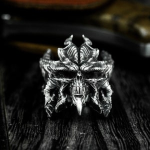 May include: A silver ring with a detailed design of a skull with horns. The ring is made of metal and has a textured surface.