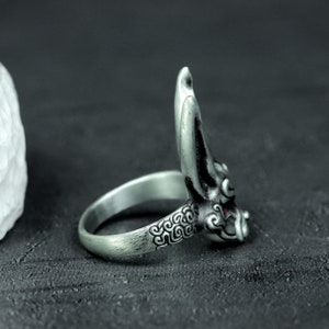 May include: Silver ring with a detailed design of a demon face with red eyes. The ring is made of metal and has a textured surface.