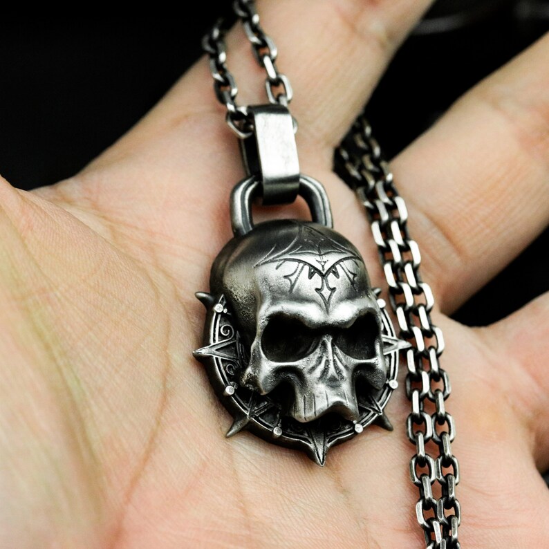 May include: A silver skull pendant necklace with a detailed design and a spiked halo. The necklace is on a silver chain.
