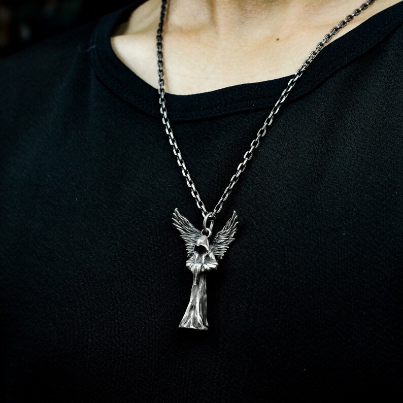 May include: A silver chain necklace with a pendant of a hooded angel with wings. The angel is standing with its arms crossed and has a detailed, textured design.