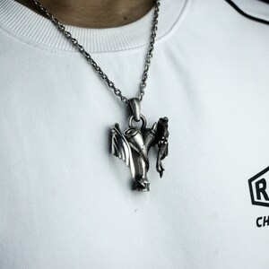 Puede incluir: Un collar de plata con un colgante que representa un &aacute;ngel y un demonio entrelazados. El colgante es detallado e intrincado, con alas, cuernos y un halo.
