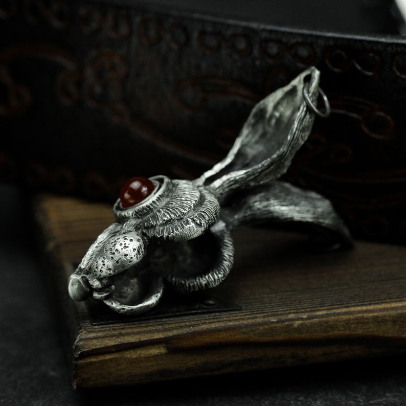 May include: A silver rabbit pendant with a red gemstone eye. The pendant has a detailed, textured surface and a loop for attaching a chain.