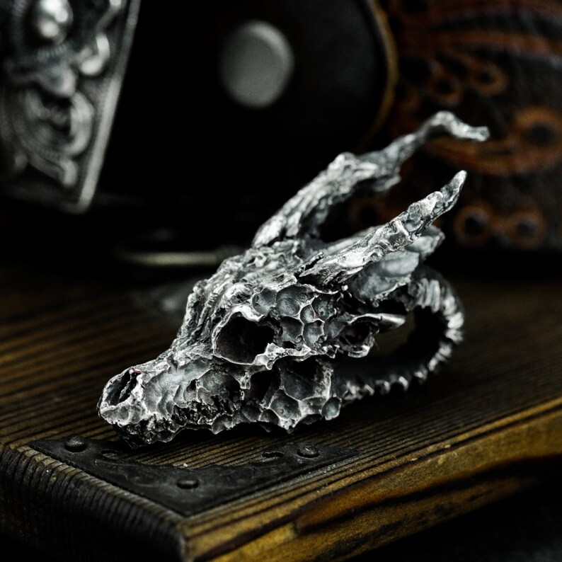 May include: A detailed silver dragon skull pendant with a textured surface. The pendant is made of metal and has a realistic design.