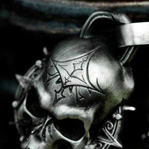 May include: A silver skull pendant with a detailed design and a spiked halo. The skull has a stylized cross etched on the forehead.
