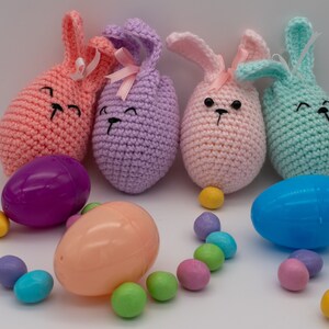 Puede incluir: Cuatro huevos de Pascua de ganchillo en forma de conejo en rosa, morado, rosa claro y azul claro. Los huevos están rodeados de huevos de chocolate cubiertos de caramelo de colores.