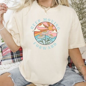 May include: A cream colored t-shirt with a graphic of a sun, clouds, and flowers in a circle. The text "Keep Moving Forward" is written around the circle.