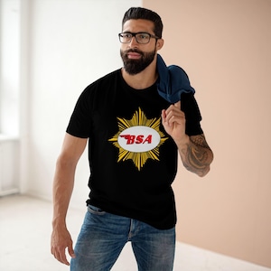 May include: Black t-shirt featuring a BSA logo. The logo is a red and white oval with the letters "BSA" inside, surrounded by a yellow burst design. The shirt is paired with blue jeans.