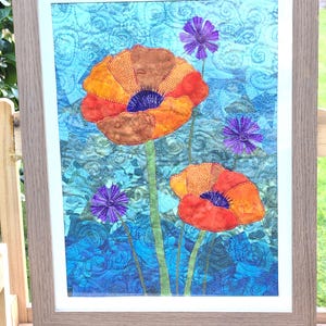 Peut inclure: Tableau textile encadré représentant des coquelicots orange et marron avec des centres et des accents violets, sur un fond floral bleu. L'œuvre est encadrée dans un cadre en bois marron clair, pour la décoration.
