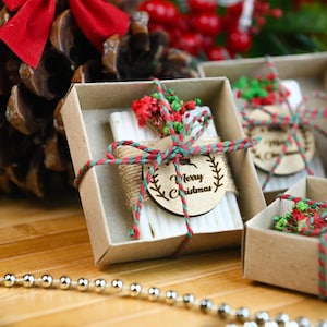 May include: Small gift boxes with a Christmas theme. Each box contains a white item wrapped with twine and a wooden tag that says "Merry Christmas". The boxes are decorated with red and green accents.
