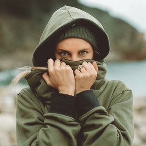 May include: A woman wearing a green hooded sweatshirt with the hood pulled up over her head. She is covering her face with her hands.