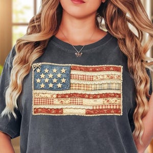 May include: Dark gray t-shirt featuring a patchwork American flag design. The flag incorporates blue, red, and cream-colored fabrics with star and stripe patterns. The shirt has short sleeves and a relaxed fit, suitable for casual wear.