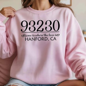 May include: A light pink sweatshirt with the text "93230 Home is where the love in Hanford, CA" in black. The sweatshirt is a crew neck style and is made of a soft material. The text is centered on the front of the sweatshirt.