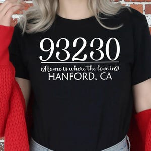 May include: Black t-shirt with the white text "93230 Home is where the love in Hanford, CA." The shirt is paired with blue jeans and a red sweater. The background is a brick wall.