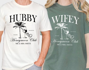 Puede incluir: Camisetas color marfil y verde musgo con las palabras "Hubby" y "Wifey" impresas. Cada camiseta presenta una palmera, una copa de vino, anillos y las palabras "Honeymoon Club" y "Mr. & Mrs. Smith".