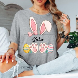 May include: A gray t-shirt featuring a bunny ears design with a pink bow and polka dots. The name "Sara" is written below the ears, with Easter eggs and bunny paw prints at the bottom. The shirt is a casual, festive design.