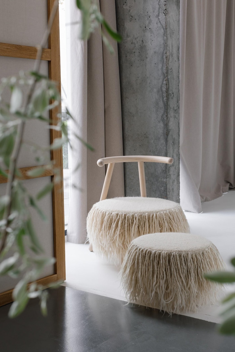 May include: A wooden chair with a fluffy white fringe seat and a matching ottoman. The chair and ottoman are in front of a concrete wall and white curtains.