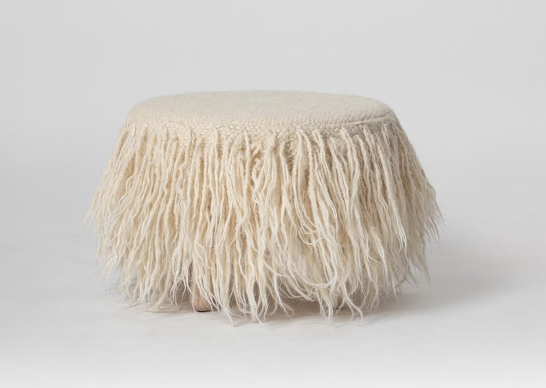 May include: A round, white, fluffy ottoman with a fringe around the top. The ottoman is sitting on a white background.