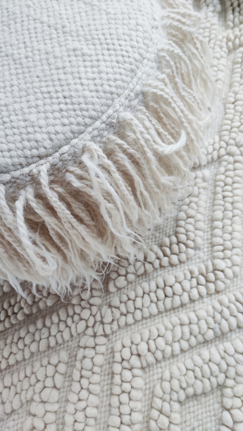 May include: A white round fluffy pillow with a fringe border. The pillow is sitting on a white textured rug.