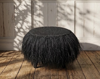 May include: A black, round ottoman with a textured top and fringe-like trim. The ottoman sits on a dark metal base and is placed on a wooden floor. The background features a white paneled wall.