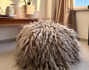 May include: A round, low-profile ottoman covered in long, shaggy, light beige fibers. The textured surface consists of dense, twisted strands of material that create a voluminous, rounded shape, resting directly on a light-colored speckled floor.