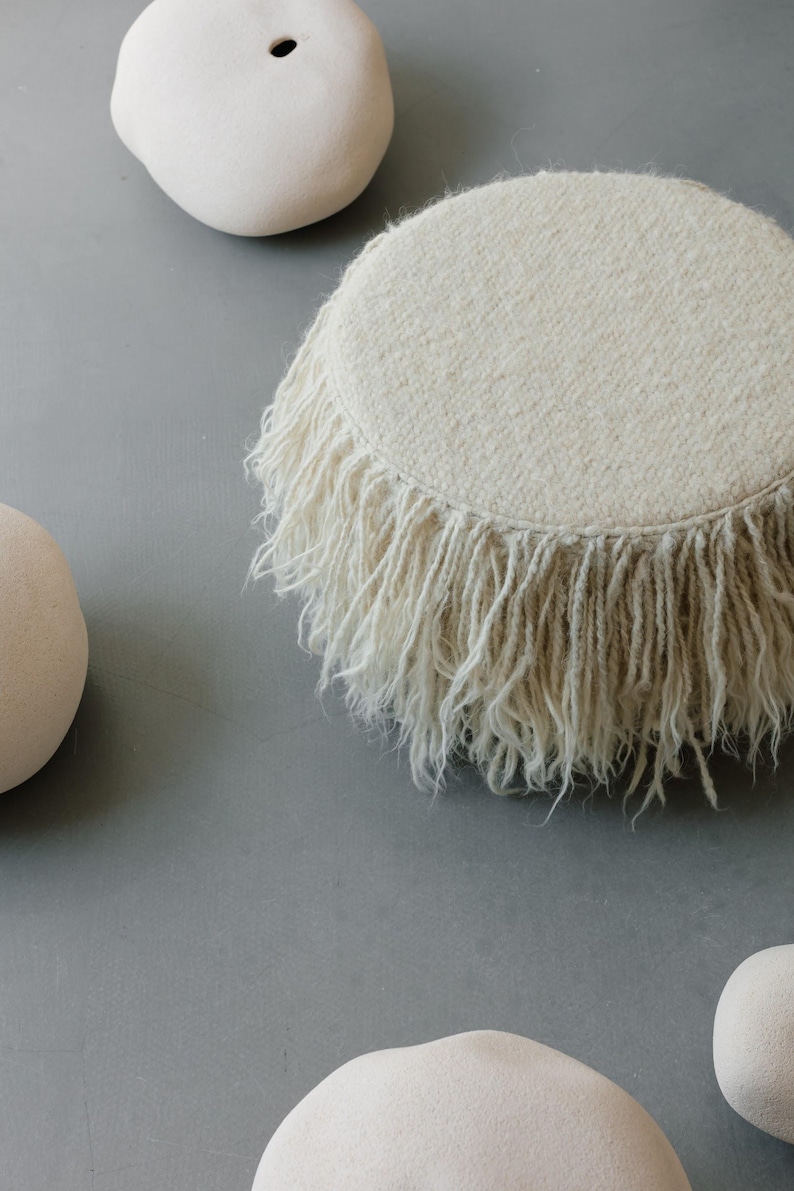 May include: A white fluffy round pouf with a fringe border sits on a gray surface with three white ceramic rocks.