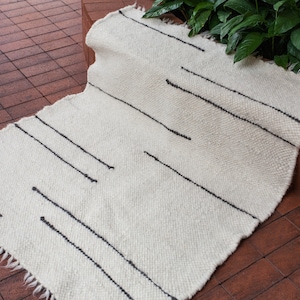 May include: Off-white woven rug with black linear accents. The rug has a rectangular shape with a fringed edge. The design features several black lines running diagonally across the surface. The rug is placed on a brick surface.