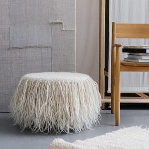 A white fluffy ottoman with a fringe around the bottom. The ottoman is sitting on a white rug in a room with a wooden chair and a wall with a textured surface.