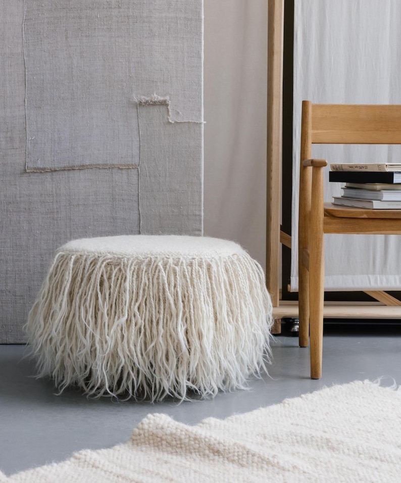 A white fluffy ottoman with a fringe around the bottom. The ottoman is sitting on a white rug in a room with a wooden chair and a wall with a textured surface.