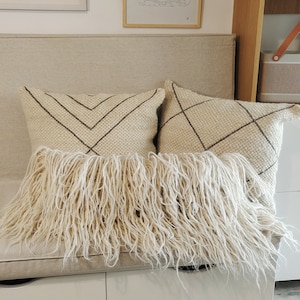 May include: Two cream-colored throw pillows with fringe trim and a geometric pattern in brown thread. The pillows are on a light beige linen bench with white drawers.