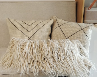 May include: Two cream-colored throw pillows with fringe trim and a geometric pattern in brown thread. The pillows are on a light beige linen bench with white drawers.
