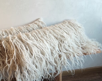 May include: Two cream-colored, shaggy pillows rest on a light wood bench. The pillows have long, textured fibers, creating a soft, inviting appearance. The bench is supported by simple wooden legs, and the background is a neutral-toned wall.
