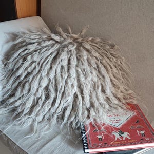 May include: A fluffy, gray, round pillow sits on a light gray couch with a red book titled "Heart Mountain" by William Grill and the words "Flying Eye Books" on the cover.
