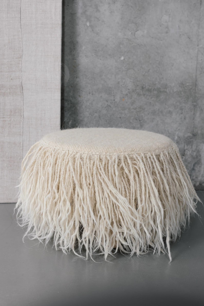 A white fluffy round ottoman with a fringe border. The ottoman is sitting on a gray surface.