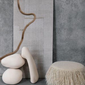 May include: A white fluffy ottoman with a fringe trim sits on a gray concrete floor.  Two white ceramic sculptures and a piece of beige fabric are in the background.