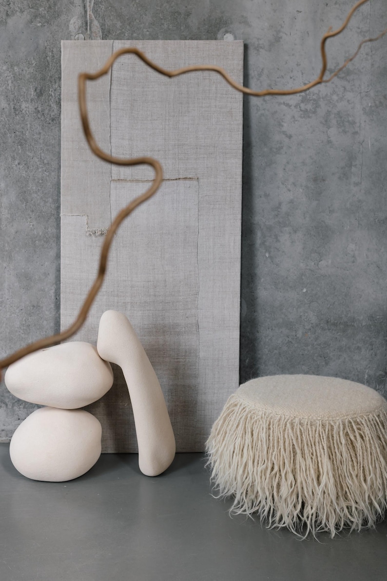 May include: A white fluffy ottoman with a fringe trim sits on a gray concrete floor.  Two white ceramic sculptures and a piece of beige fabric are in the background.