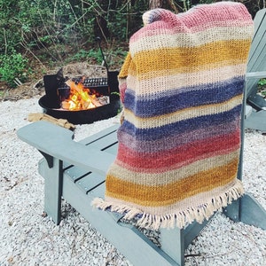 Puede incluir: Una manta de punto a rayas con flecos en los bordes, drapeada sobre una silla de madera frente a una chimenea. La silla es de madera gris claro y está sentada sobre una cama de grava blanca.
