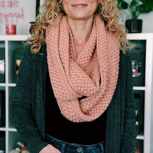 Könnte beinhalten: Eine Frau trägt einen strukturierten Rundschal in Staubrosa. Sie trägt außerdem eine dunkelgraue Strickjacke, ein schwarzes Oberteil und blaue Jeans. Der Schal scheint handgestrickt oder gehäkelt zu sein.