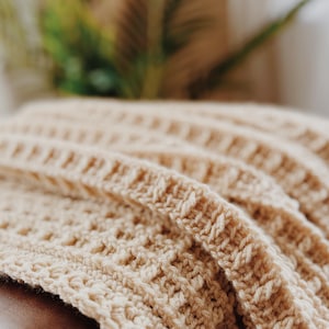 May include: A close-up of a beige crocheted throw blanket draped over a brown leather sofa.